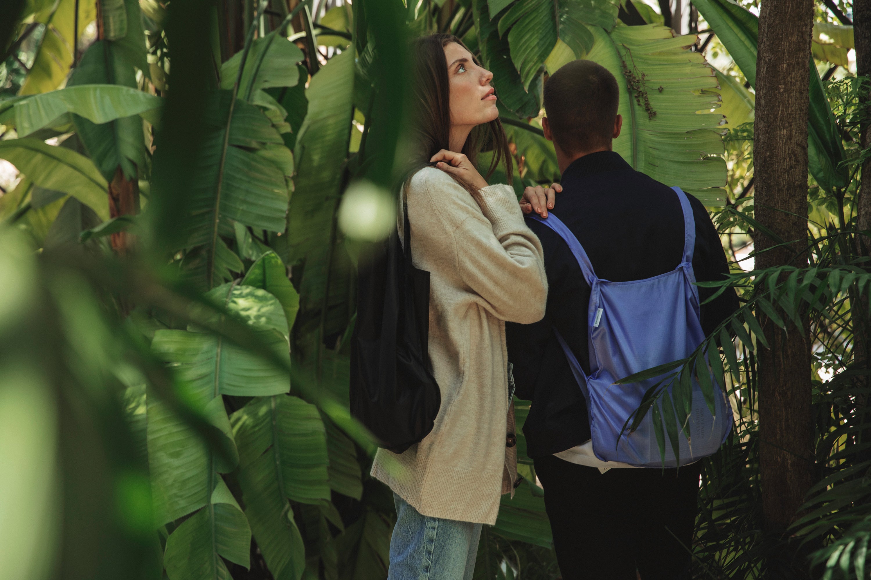 Two models wearing cornflower purple and black Notabags while visiting a botanic garden, surrounded by a variety of plants, showcasing the bags' stylish design.