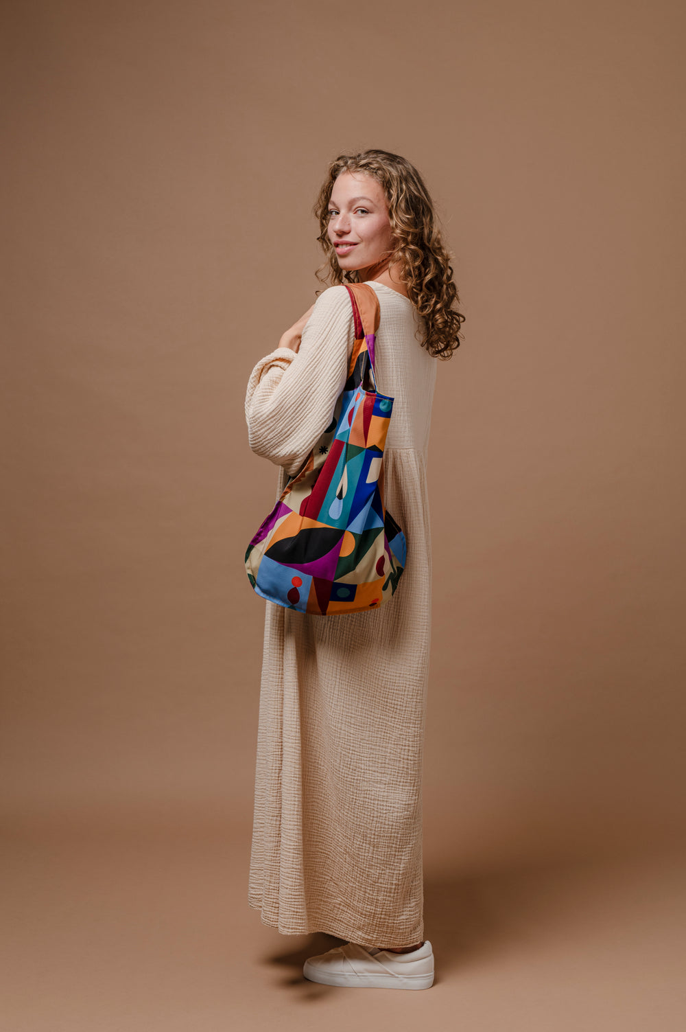 A woman in a long cream knit dress stands against a solid tan studio background, holding a colorful geometric print shoulder bag.