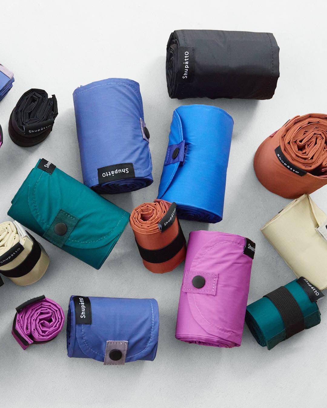 A collection of rolled camping sleeping bags in various colors, including blue, purple, teal, black, orange, and beige, arranged on a light gray surface. Each bag features a small black label with the brand name 'Soufflo' printed in white.