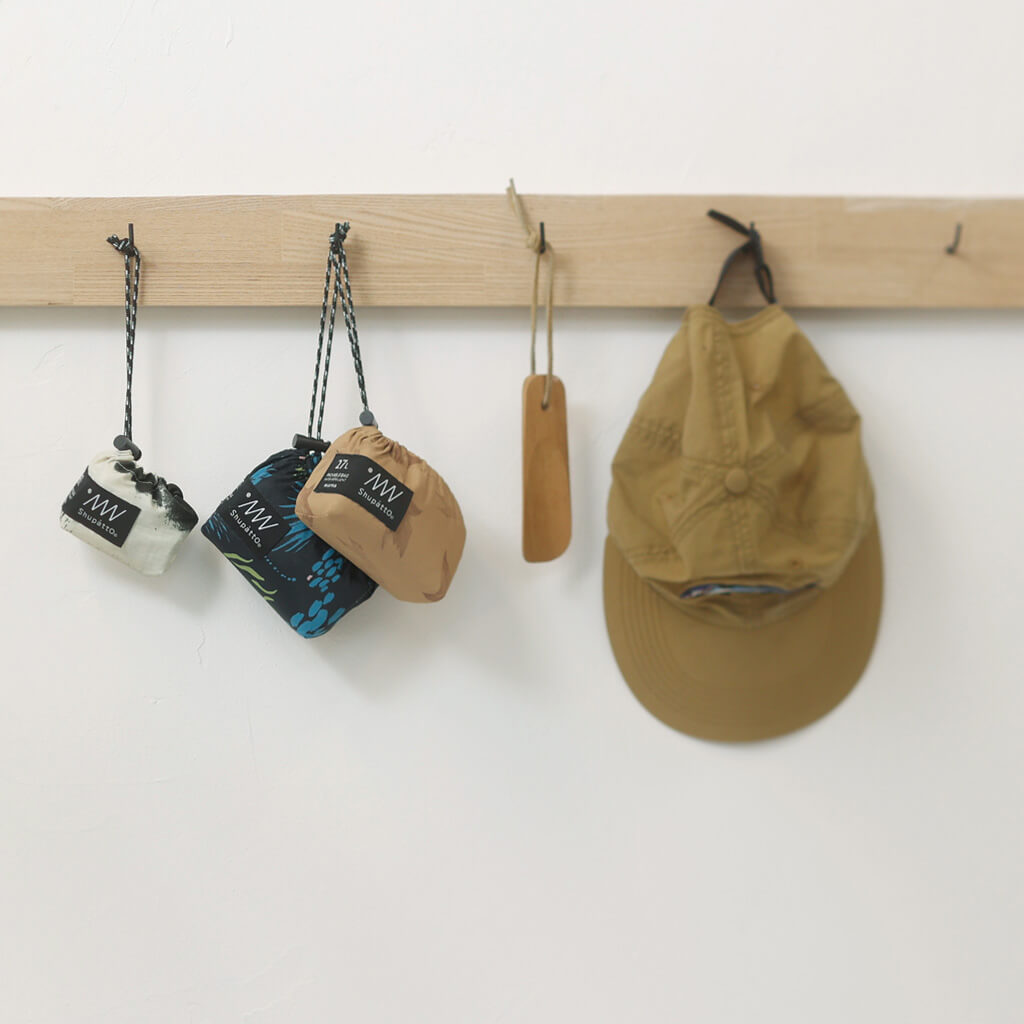 Three small Shupatto packable bags and a brown cap hanging on a wooden rack against a white wall.