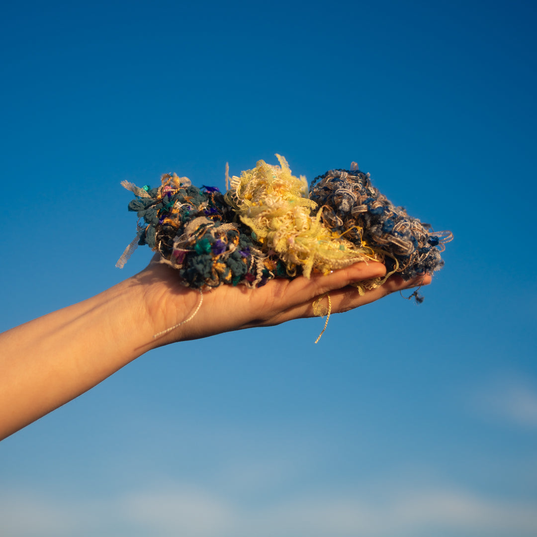 Three handmade crochet scrunchies: a vibrant blue and orange one, a cheerful yellow one with confetti decorations, and a delicate blue one made of intertwined threads, all swaying gently in the breeze.