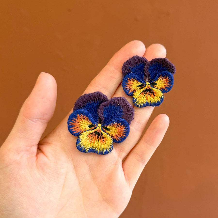 Hand holding two embroidered pansy flowers against a brown background