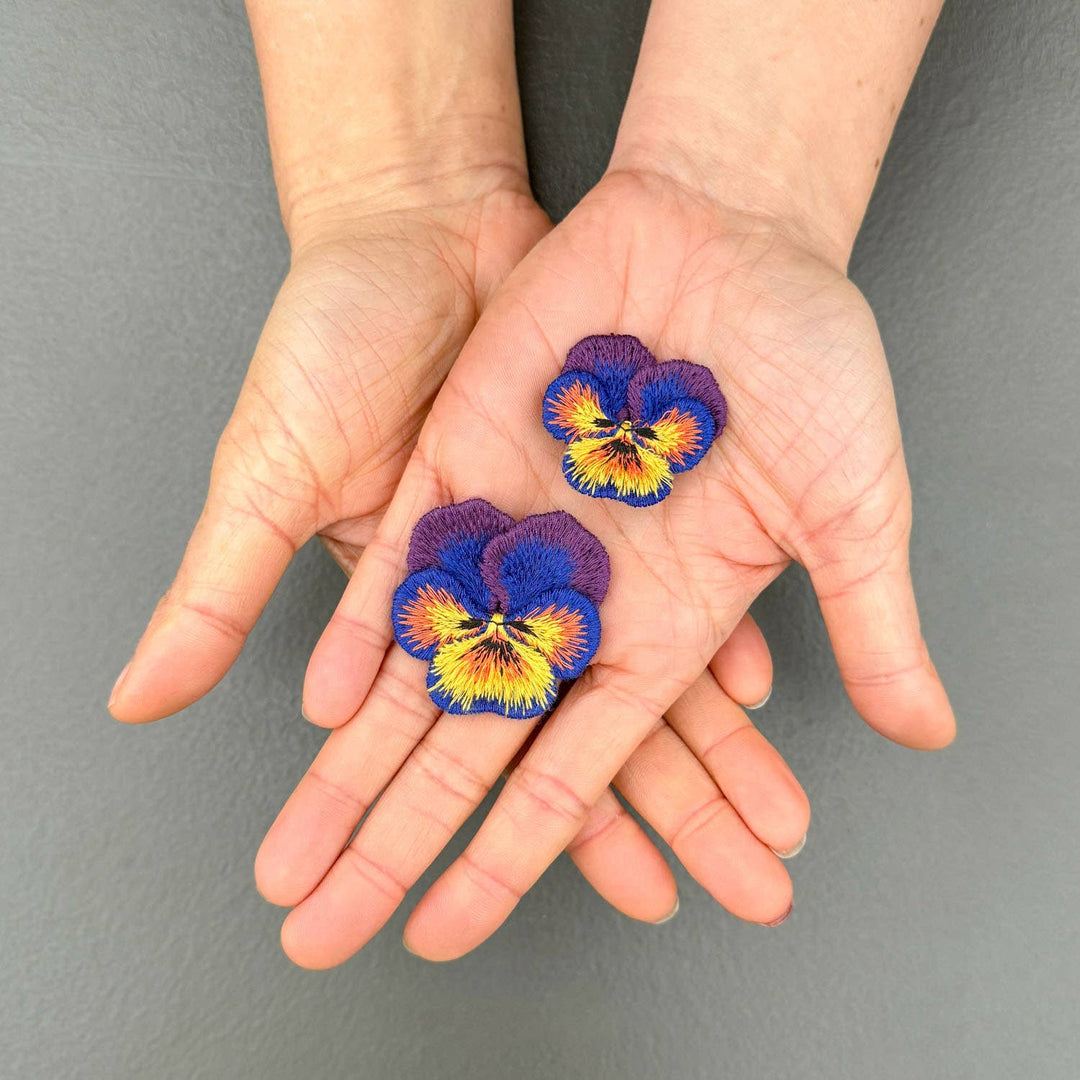 Two hands holding colorful pansies embroidered brooches against a gray background
