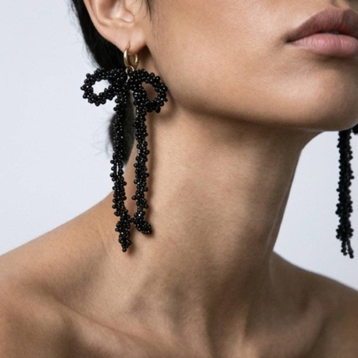 Black beaded bow earrings worn by a person on a neutral background